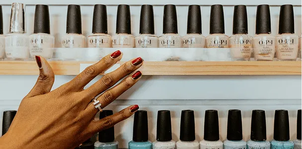 A hand with a deep red manicure in front of shelves of premium nail polish
